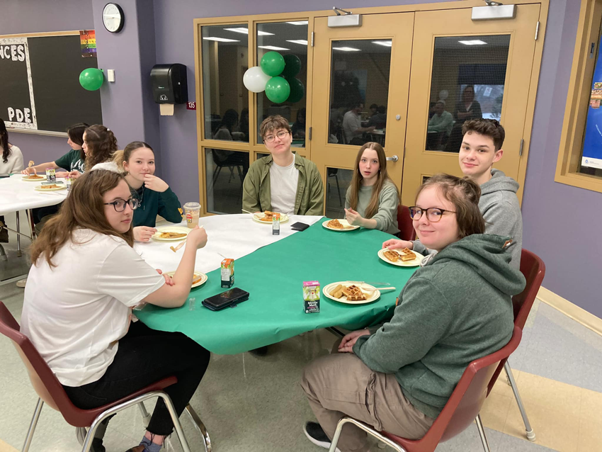 Des élèves mange un repas sur une table avec une nappe blanche et vert représentant le drapeau de la francophonie ontarienne