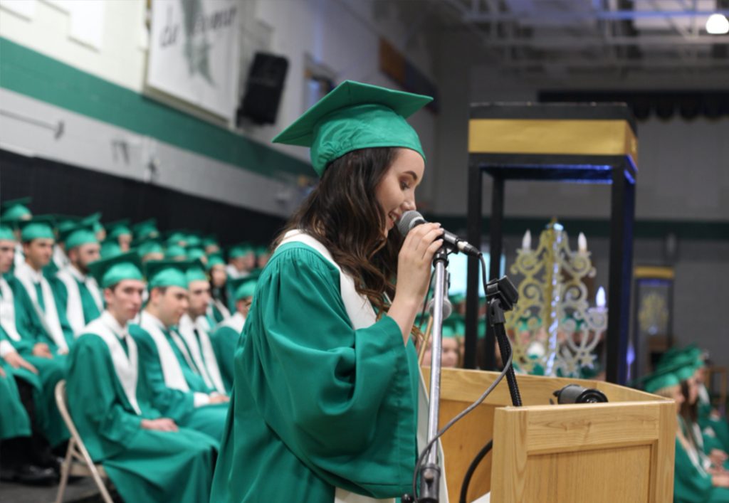 L'image montre une personne portant une robe verte et un bonnet parlant dans un microphone lors d'une cérémonie de remise des diplômes du secondaire.