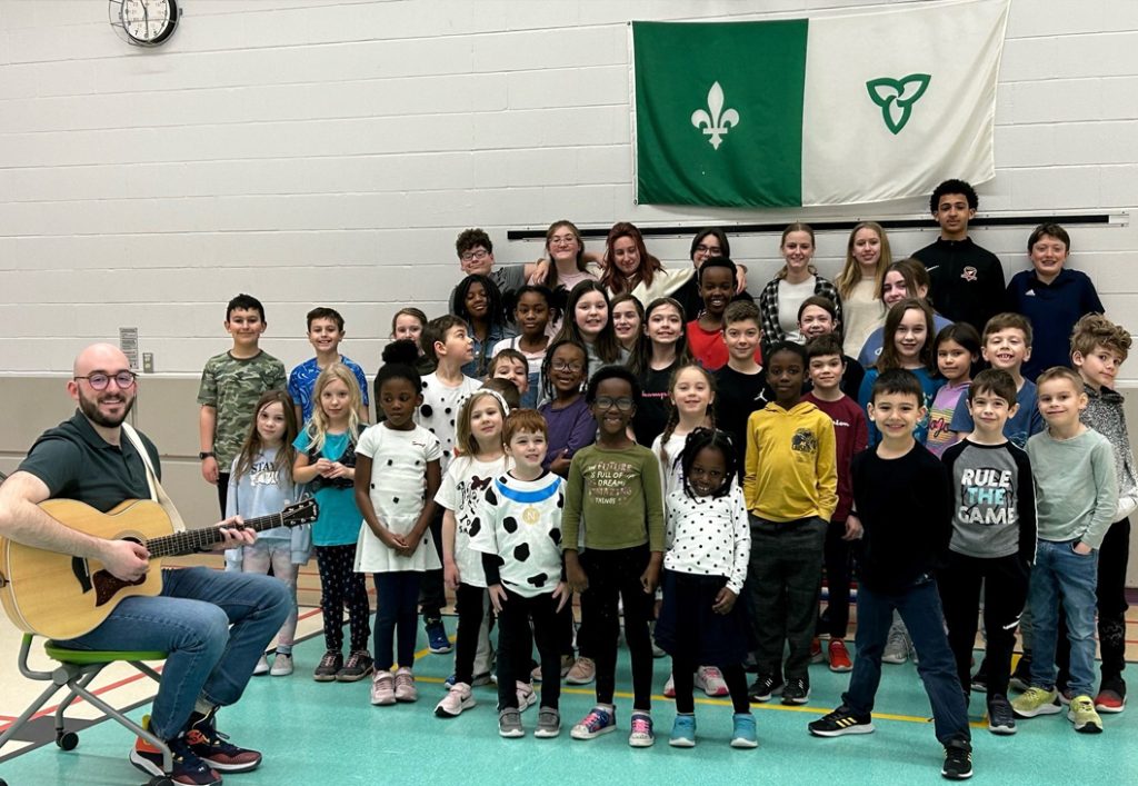 Un groupe d'enfants choristes pose pour une photo avec leur enseignant assis sur un tabouret à roulettes, tenant une guitare prêt à commencer à jouer.