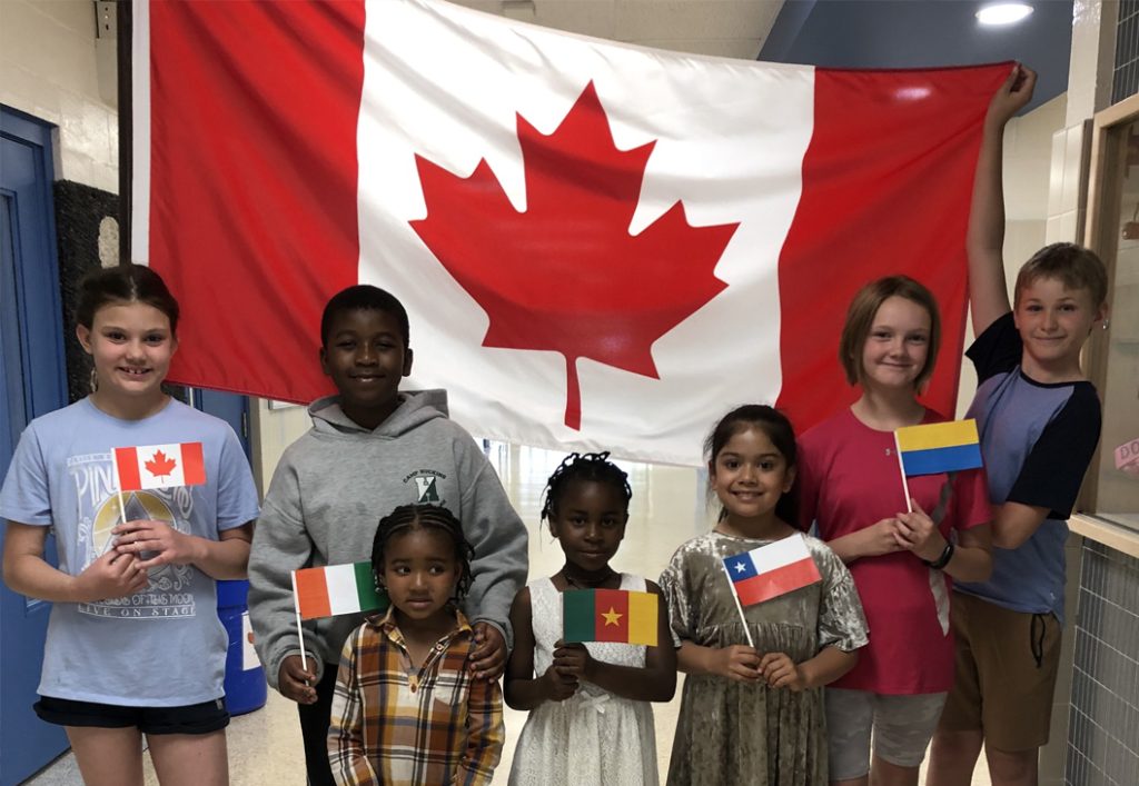 Des enfants de différentes nationalités tiennent leur drapeau national en se tenant derrière un grand drapeau du Canada.