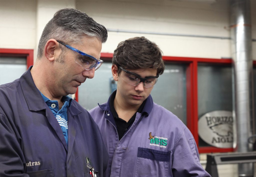 Deux hommes debout dans un atelier, portant des lunettes et des combinaisons, regardant quelque chose hors de l'écran.