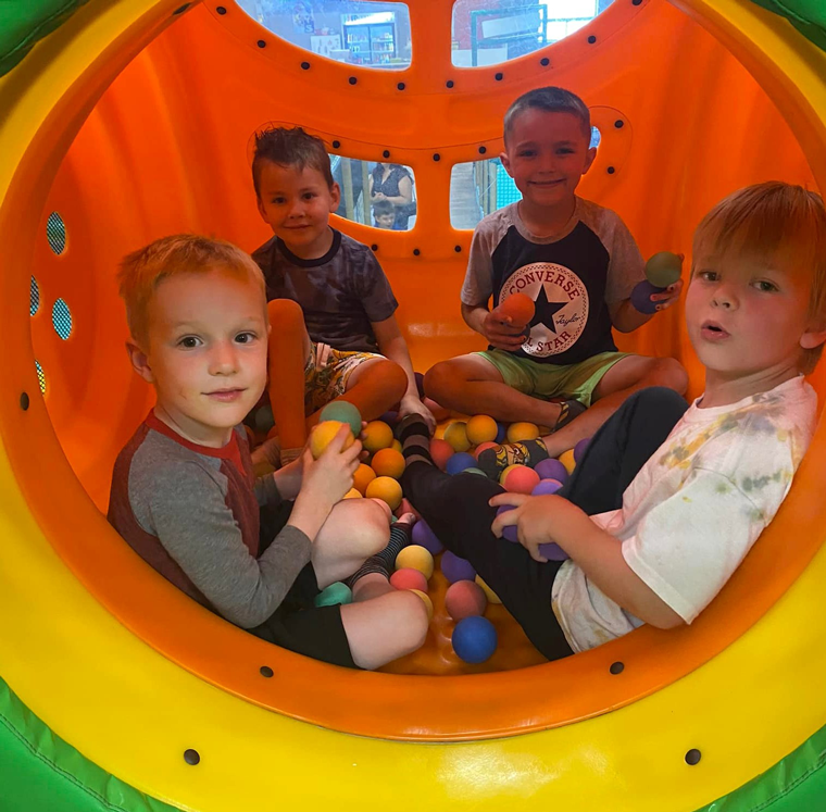 Quatre enfants assis avec joie dans une fosse à balles vibrante, entourés d'un éventail de balles colorées.